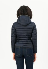 Marineblauwe pufferjas met een getailleerd silhouet, horizontaal gestikt ontwerp en een capuchon. Voorzien van een gouden accent langs de kraag.