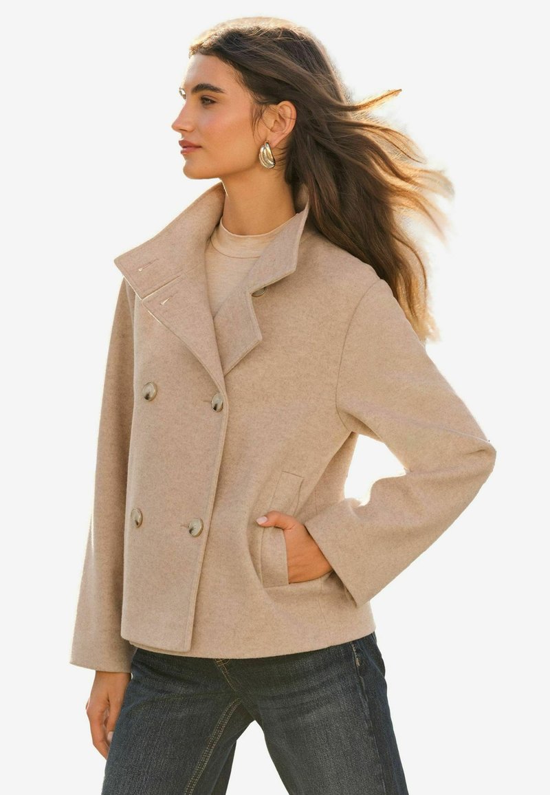 Beige dvojradový vlnený kabát s klopovým golierom, dvoma prednými vreckami a striebornými gombíkmi. Modelka pózuje s rukou vo vrecku a voľnými vlasmi.