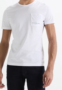 Vit bomull t-shirt med korta ärmar, rundhalsringning och en vänster bröstficka med "Calvin Klein Jeans" logotyp i svart text.