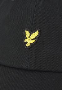 Casquette noire en tissu texturé, arborant un logo d'aigle brodé jaune sur le devant, avec une couronne structurée et une visière incurvée.