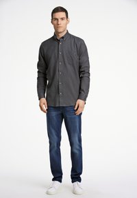 Camicia button-up grigia scura con tasca sul petto, abbinata a jeans blu e sneakers bianche. La camicia ha bottoni in legno e maniche lunghe.