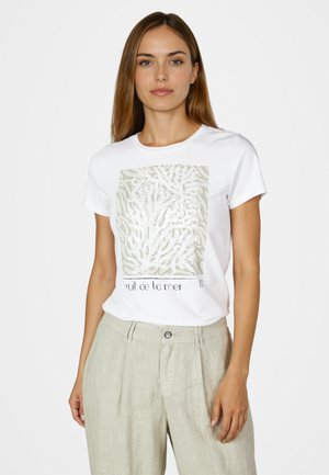 Junge Frau trägt weißes T-Shirt mit abstraktem Korallendesign und beigefarbenen hoch taillierten Hosen vor einfachem Hintergrund.