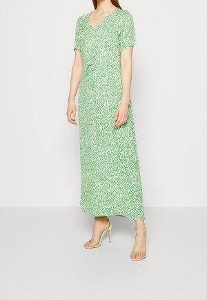 Frau trägt ein knöchellanges Kleid mit grün-weißem Muster, kurzen Ärmeln und grüne, Riemchen-Sandaletten mit hohen Absätzen.