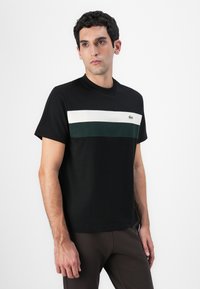Schwarzes Baumwoll-T-Shirt mit einem horizontalen Farbblockdesign in Weiß und Grün. Gerippter Rundhalsausschnitt und Lacoste-Logo-Akzent auf der Brust.