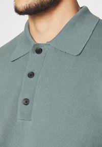 Polo vert menthe texturé avec un col à revers et trois boutons noirs. Vue en gros plan mettant en valeur les détails du tissu et les coutures.