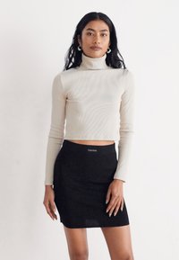 Krém színű, bordázott, magas nyakú crop top, amihez egy szűk fekete mini szoknya társul. A top hosszú ujjú, míg a szoknya derékrészén logó található.