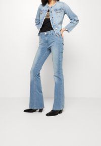 Mujer con jeans acampanados de color azul claro, chaqueta de mezclilla corta con botones plateados, botas de tobillo negras con punta afilada y blusa de estampado de leopardo.