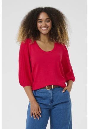 Red knit top with a scoop neckline and three-quarter puff sleeves, textured detailing on sleeves, paired with high-waisted blue denim.