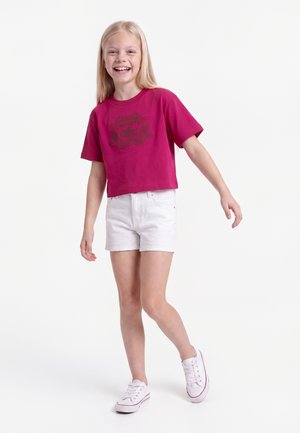 Lächelndes junges Mädchen steht mit leicht ausgestreckten Armen, trägt ein pinkes Crop-Top, weiße Shorts und weiße Turnschuhe vor weißem Hintergrund.
