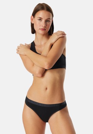 Zwarte sportbeha en bijpassende bikinionderzijde van gladde stof. De tailleband heeft een reliëfmerk. Model poseert met de armen gekruist.