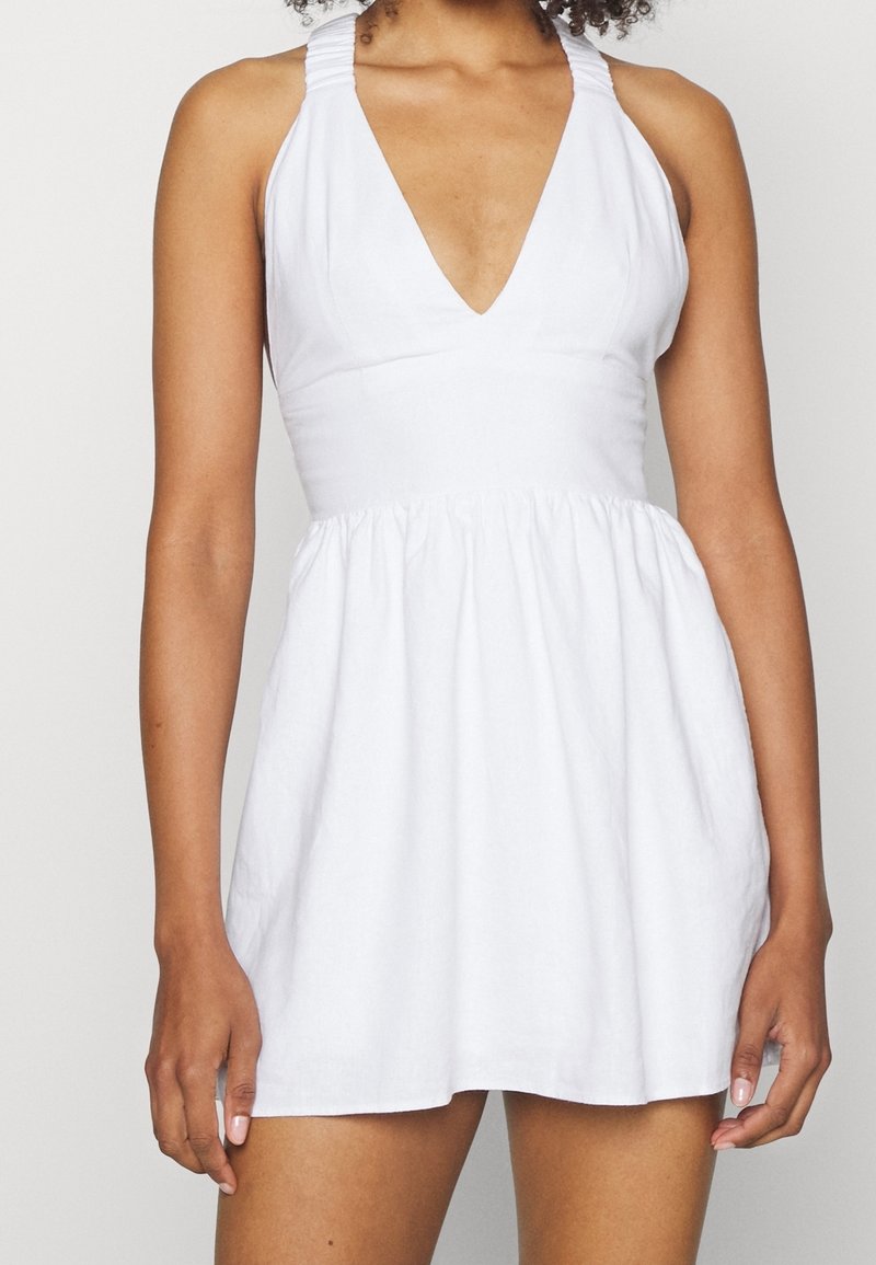 Bílé halter šaty s hlubokým výstřihem do V, nařaseným pasem a texturovanou látkou, s rozšířenou sukní a úzkými ramínky.
