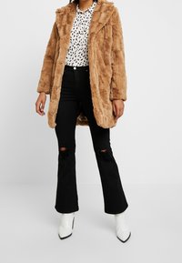 Beige pälsjacka med texturerad design, bärs över en vit skjorta med mönster och svarta utsvängda jeans. Vita spetsiga ankelboots.