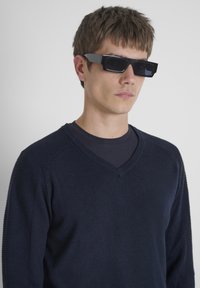 Zwarte rechthoekige zonnebril met donkere lenzen. Het montuur is dik en hoekig. Het model draagt een marineblauwe V-hals trui met ribbel-details.