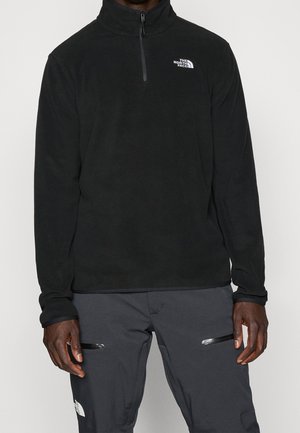 Mężczyzna w czarnym polarze z zamkiem do ćwiartki i białym logo North Face oraz ciemnoszarych spodniach z zamykanymi kieszeniami na udach, stojący na jednolitym tle.