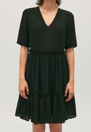 Robe noire mi-longue, manches courtes, encolure en V, avec des détails froncés volantés à la taille et à l'ourlet, portée par une personne debout.