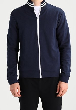 Mann trägt eine marineblaue Jacke mit Reißverschluss, weiß gestreiftem, geripptem Kragen und schwarze Hose, steht vor einem schlichten, hellen Hintergrund.