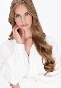 Camicia bianca con maniche strutturate, con dettagli di bottoni. Il modello ha capelli lunghi e ondulati. Lo sfondo bianco semplice enfatizza l'attenzione sul capo.
