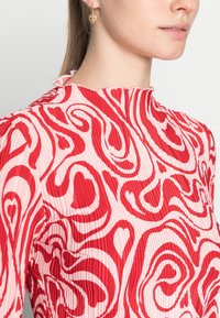 Röd och rosa plisserad topp med ett snurrat hjärtmönster, med hög krage och volangkant. Guldörhängen i form av hoops kompletterar designen.