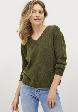 Olivgrüner, locker sitzender V-Ausschnitt-Pullover mit langen Ärmeln und gerippten Bündchen, kombiniert mit hellblauen, engen Jeans. Weiche Textur.