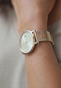 Goldfarbene Armbanduhr mit einem silbernen Mesh-Armband, rundem Zifferblatt, hellcremfarbener Ziffernblatt und subtilen Branding-Details.