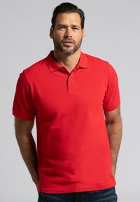 Czerwona koszulka polo wykonana z bawełny, z klasycznym kołnierzykiem, zapięciem na dwa guziki i krótkimi rękawami, noszona przez mężczyznę na neutralnym tle.