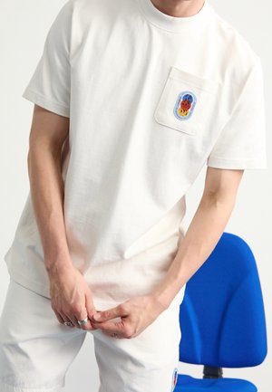 Homme portant un t-shirt blanc et un short avec des patchs colorés, debout devant une chaise de bureau bleue, les mains jointes près de la taille.