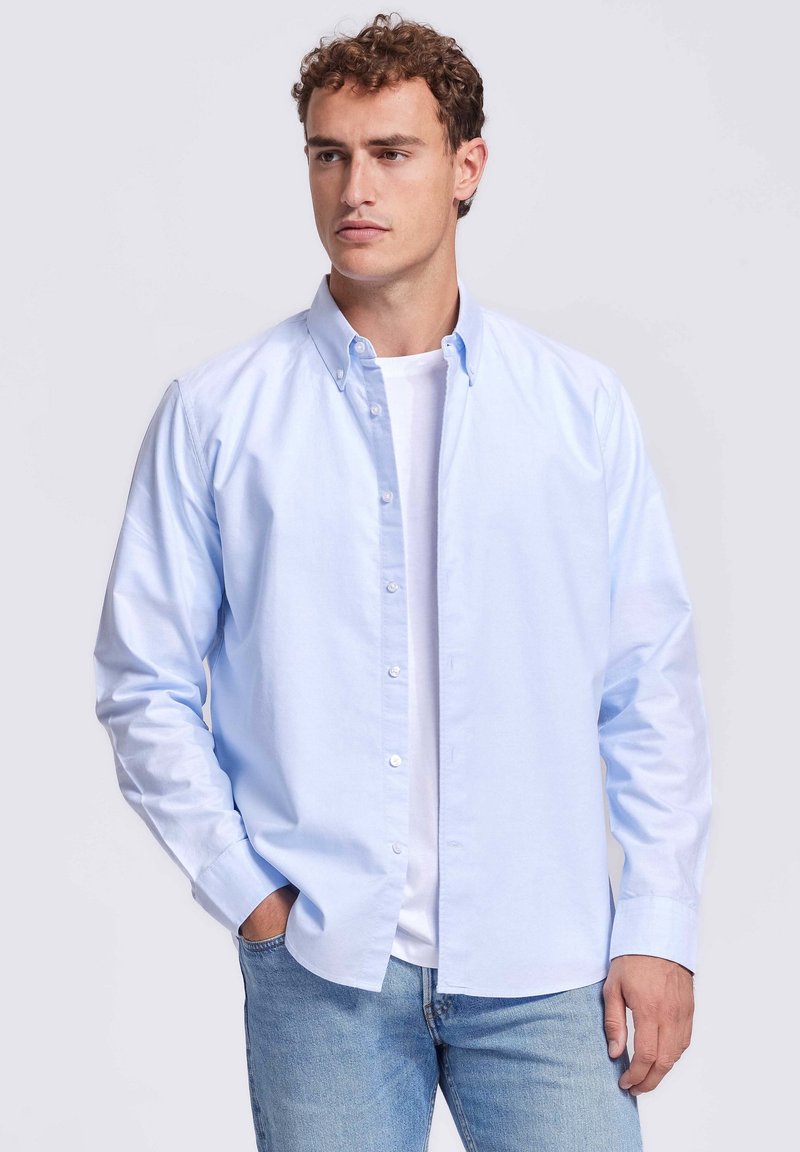 Man met krullend haar, gekleed in een lichtblauwe overhemd met knopen over een wit T-shirt, staand met één hand in zijn zak tegen een witte achtergrond.