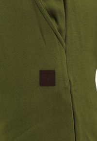 Tkanina w oliwkowym kolorze o gładkiej fakturze, z brązową skórzaną metką z napisem "KEY LARGO". Prosty, dopasowany fason bez wzorów.