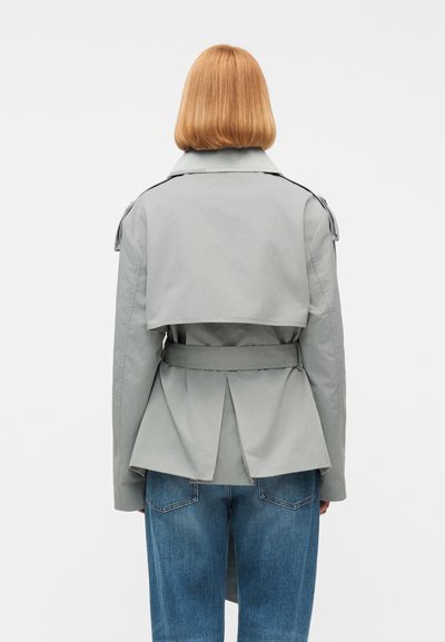 Άτομο με ίσια κόκκινα μαλλιά, φοράει ένα ανοιχτό γκρι καμπαρντίνα με ζώνη και μπλε τζιν, φαίνεται από την πλάτη σε απλό λευκό φόντο.