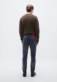 Brun ribbestrikket genser, blå jeans med rødt merke på linningen og svarte sko. Bildet viser bakfra av antrekket.