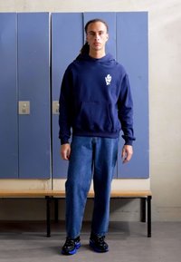 Marineblå hettegenser med logo på brystet, parret med løse dongeribukser og svarte joggesko med blå detaljer. Bakgrunnen viser skap.