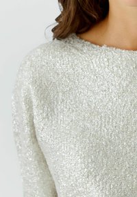 Gros plan d'une personne portant un pull texturé gris clair avec un tissu doux et nuageux sur un fond uni.