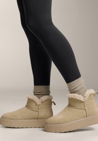 Barna színű velúr bokacsizma szőrmés díszítéssel, texturált talppal és hurkokkal a könnyű felvételhez, fekete leggingssel és krémszínű bordázott zoknival párosítva.