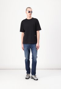 T-shirt noire à manches courtes avec un col rond, associé à un jean bleu et des baskets grises. Les accessoires comprennent des lunettes de soleil réfléchissantes.