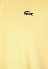 Polo de couleur jaune pâle, fabriqué en tissu texturé. Il présente un petit logo crocodile brodé en vert et rouge sur la poitrine.