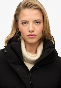 Chaqueta de plumas negra con un cuello alto y cierre de cremallera, combinada con un suéter de cuello alto de punto en un tono beige claro. Texturas suaves y un diseño elegante.