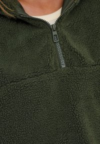 Sudadera de forro polar en verde oscuro con un cuello de media cremallera. La tela texturizada y suave presenta una cremallera lisa y una costura sutil a lo largo del pecho.