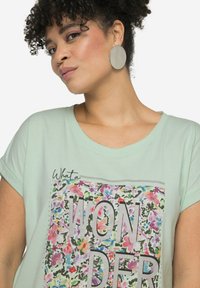Camiseta de algodón verde menta con mangas cortas, que presenta un gráfico floral con la palabra "WONDER" en letras gruesas. Acentos plateados completan el diseño.