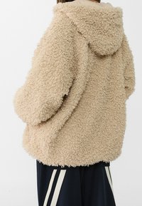 Beige, fuzzy, hooded jacket made of textured fabric with a loose fit. Features dropped shoulders and a rounded hem.