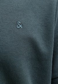 Teksturowana, ciemnozielona tkanina w wzór kraty. Zawiera mały, wyszywany symbol ampersandu w dopasowanym kolorze.