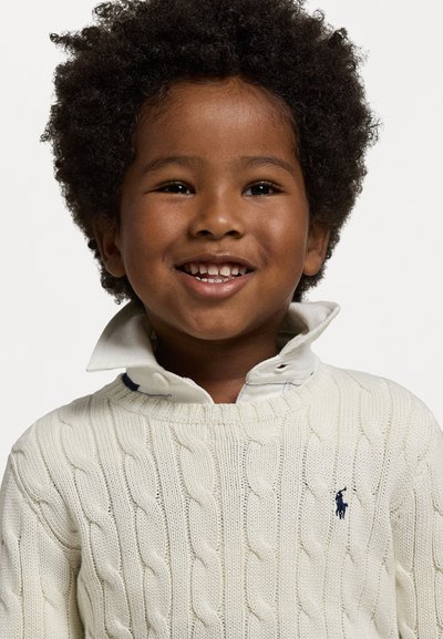 Krämfärgad kabelstickad tröja med ribbmönster och marinblå logotyp. Lagertyp krage med vit och blå ruta. Har en rund halsringning och långa ärmar.