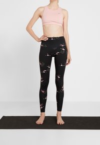 Jasnoróżowy stanik sportowy i czarne legginsy z różowymi wzorami flamingów. Materiał wydaje się elastyczny i gładki w dotyku. Modelka stoi bosą stopą na macie.