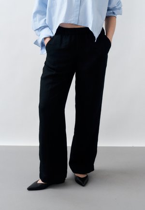 Osoba ubrana w luźne czarne spodnie, jasnoniebieską skróconą koszulę z podwiniętymi rękawami, z rękami w kieszeniach, stojąca na szarej podłodze w czarnych butach z ostrym noskiem.