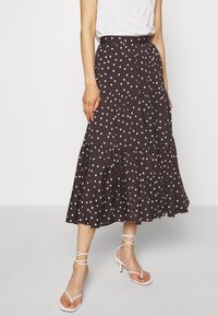 Bruine rok met witte stippen en een gelaagd ontwerp, voorzien van een comfortabele tailleband en een vloeiende zoom. Gecombineerd met witte sandalen met bandjes.