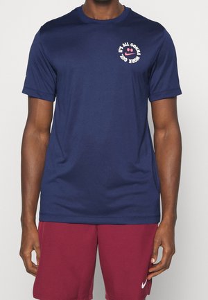 Homme portant une chemise à manches courtes bleu marine avec le texte « Ça va bien se passer » et un smiley, associée à un short de sport bordeaux avec un logo blanc.