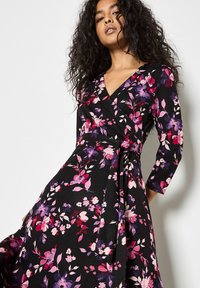 Vestido envolvente de manga larga en tejido negro con un patrón floral en tonos de rosa y púrpura, con una cintura ajustada y falda fluida.
