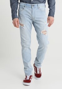Jeans de denim azul claro con detalles desgastados en la parte frontal, de corte ajustado y cinco bolsillos, combinados con zapatillas rojas y una camisa oscura de lunares.