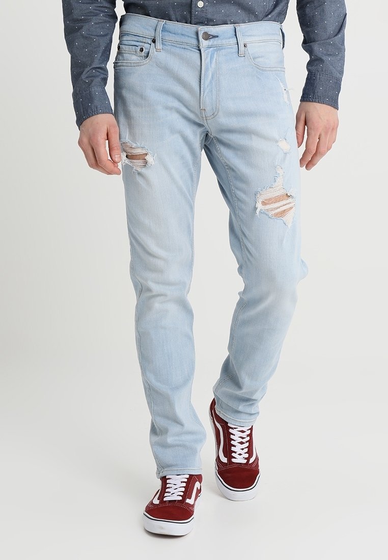 Jeans de denim azul claro con detalles desgastados en la parte frontal, de corte ajustado y cinco bolsillos, combinados con zapatillas rojas y una camisa oscura de lunares.