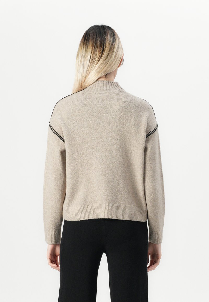 Person mit langen blonden Haaren, die einen beigen Strickpullover mit schwarzer Nahtverzierung an den Schultern und schwarze Hosen trägt und mit dem Rücken zur Kamera steht.