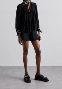 Blouse noire plissée avec un col contrastant, associée à un short noir. Le mannequin porte des mocassins noirs brillants avec une semelle épaisse.
